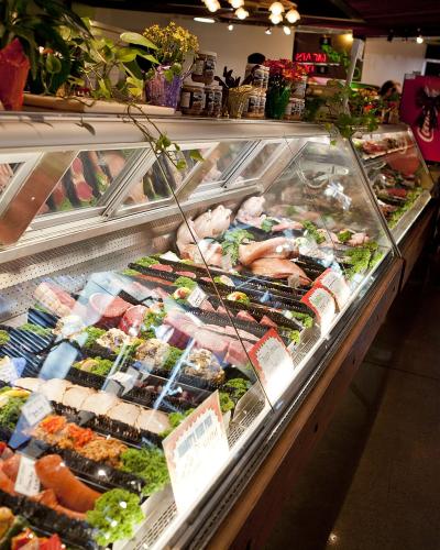 An indoor market scene showing a long refrigerated display case filled with packaged meats, sausages, and prepared foods, with shoppers walking along the aisle.