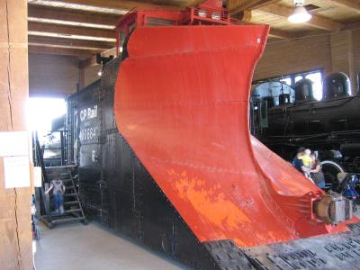 A large red and black CP Rail snowplow (number 400884) displayed indoors on tracks, with a steam locomotive visible in the background and visitors present.