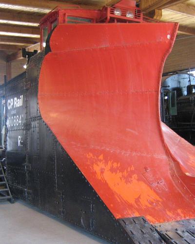 A large red and black CP Rail snowplow (number 400884) displayed indoors on tracks, with a steam locomotive visible in the background and visitors present.