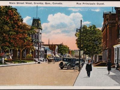 A vintage postcard depicting a wide, tree-lined commercial street with early 20th-century automobiles, pedestrians, and a prominent clock tower building on the left side.