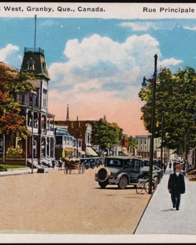 A vintage postcard depicting a wide, tree-lined commercial street with early 20th-century automobiles, pedestrians, and a prominent clock tower building on the left side.