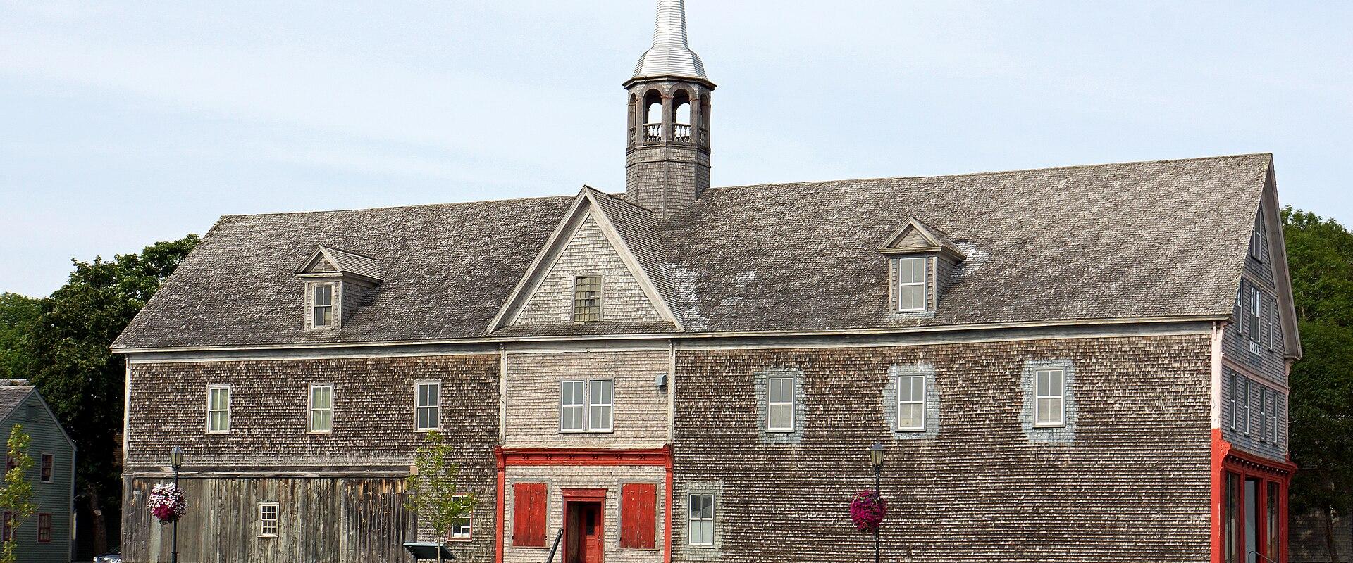 A large, historic wooden building with a shingled exterior, a central cupola with a spire, and red-painted trim, situated on a grassy waterfront with a stone seawall.