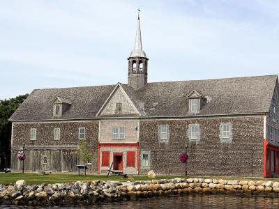 A large, historic wooden building with a shingled exterior, a central cupola with a spire, and red-painted trim, situated on a grassy waterfront with a stone seawall.