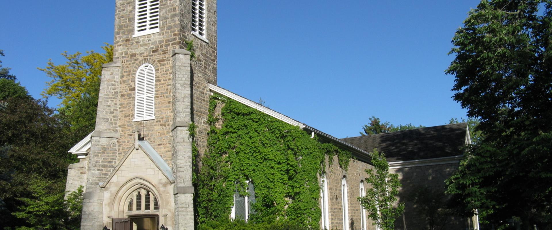 A stone church with a square tower featuring white louvered windows and a side wall heavily covered in green ivy, set against a blue sky with surrounding trees and a paved path.