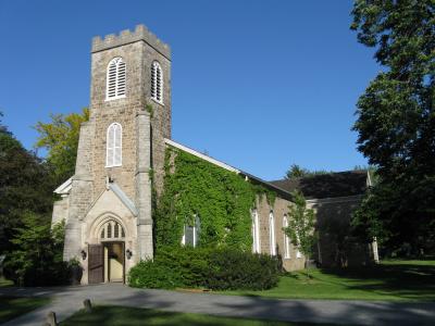 A stone church with a square tower featuring white louvered windows and a side wall heavily covered in green ivy, set against a blue sky with surrounding trees and a paved path.