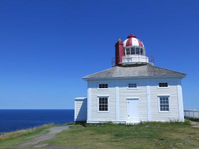 A white wooden lighthouse keeper's house with a red and white lantern room and chimney, situated on a grassy cliff edge overlooking the ocean under a clear blue sky.