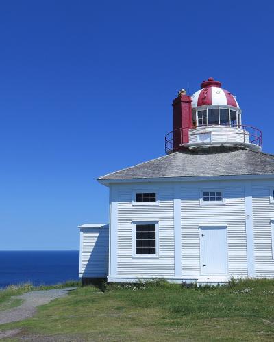 A white wooden lighthouse keeper's house with a red and white lantern room and chimney, situated on a grassy cliff edge overlooking the ocean under a clear blue sky.
