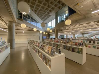 The image shows the interior of a modern library with white display tables, rows of books, large spherical pendant lights, and a high concrete ceiling with a coffered design.