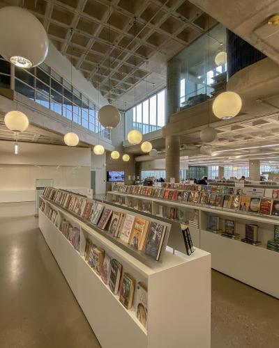 The image shows the interior of a modern library with white display tables, rows of books, large spherical pendant lights, and a high concrete ceiling with a coffered design.