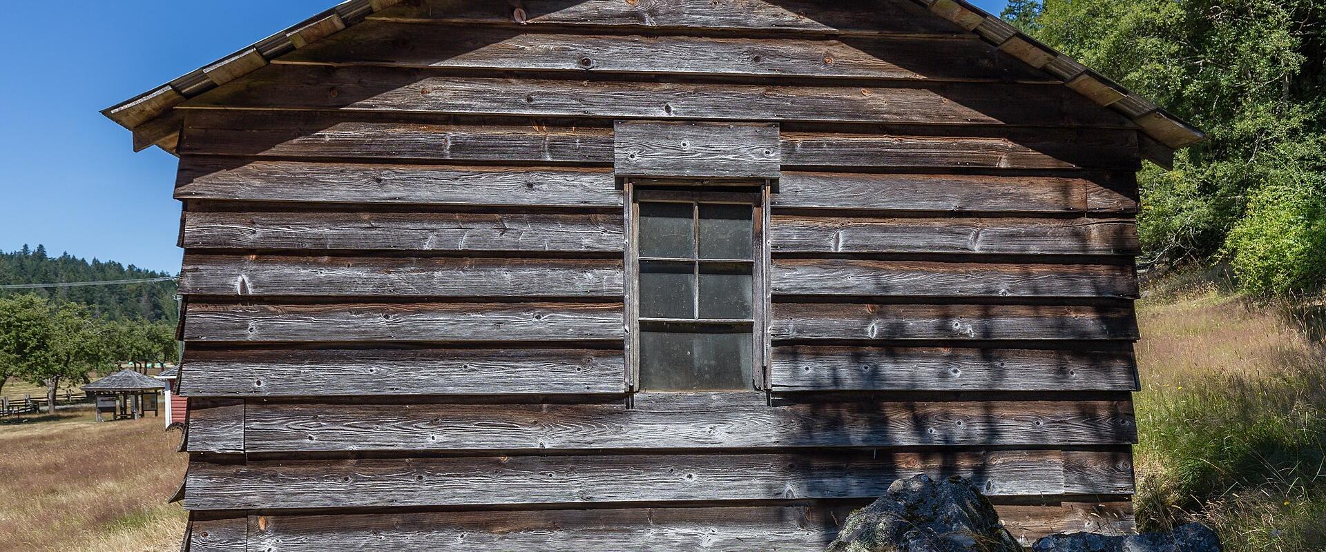 A weathered wooden log cabin with a single window, situated in a grassy field with trees and a blue sky in the background.