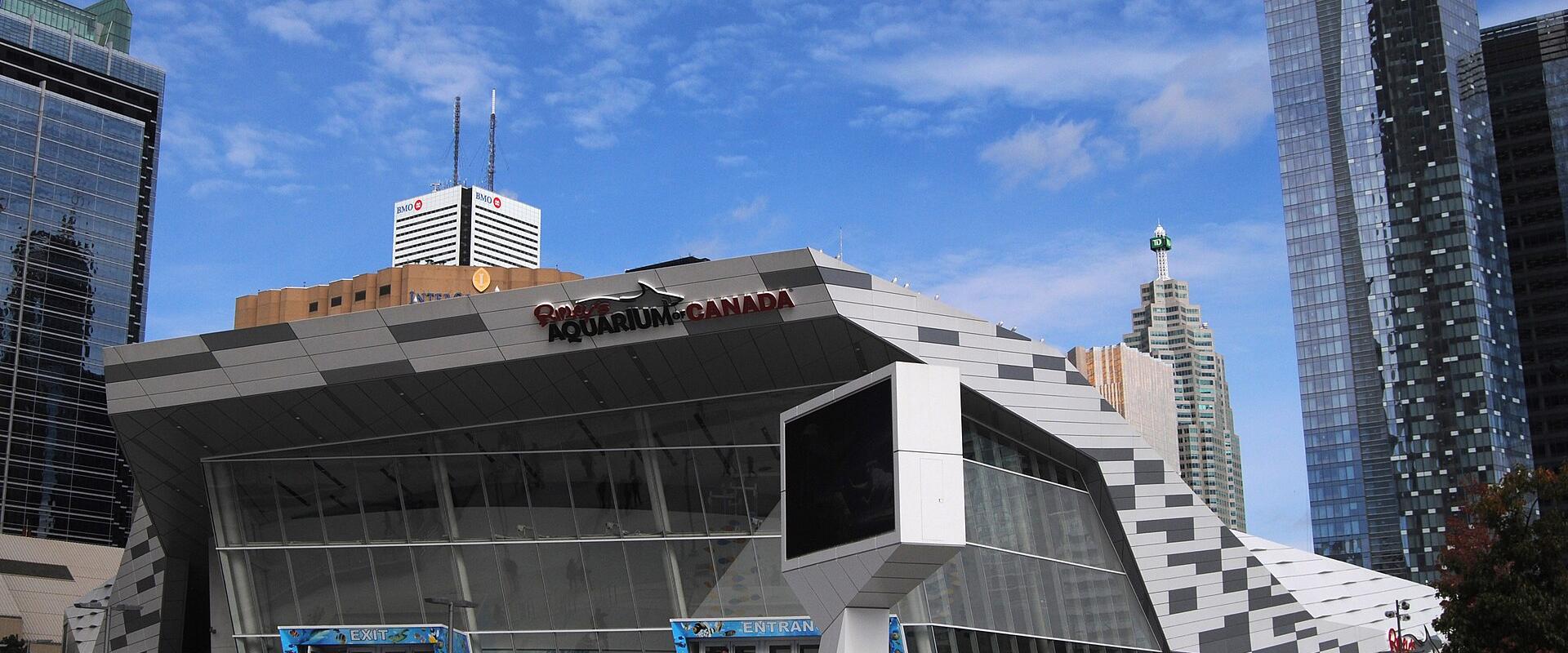 The image shows the exterior of a modern building with a large sign reading "Ripley's AQUARIUM CANADA" above the entrance, with people walking on the steps and surrounding plaza, and tall skyscrapers in the background including the CN Tower.