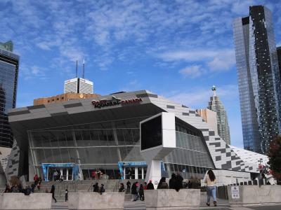 The image shows the exterior of a modern building with a large sign reading "Ripley's AQUARIUM CANADA" above the entrance, with people walking on the steps and surrounding plaza, and tall skyscrapers in the background including the CN Tower.