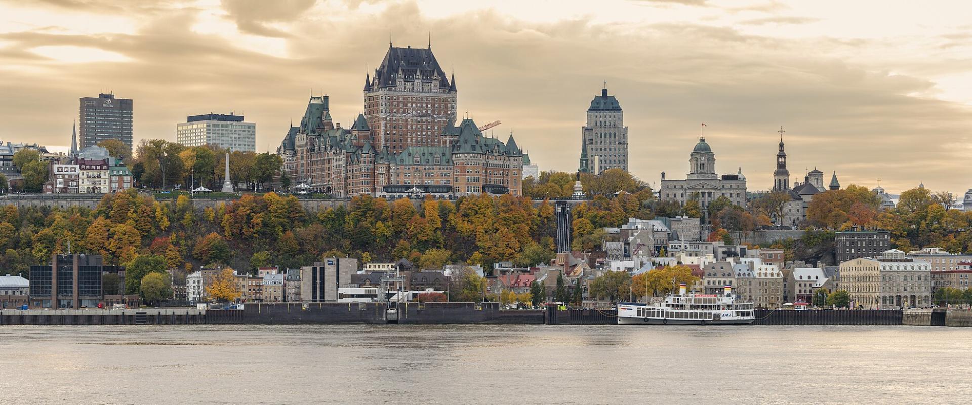 The Château Frontenac towers over Old Quebec's fortified clifftop skyline above the St. Lawrence River, with autumn foliage, the Lower Town waterfront, and a ferry at the dock as seen from Lévis.