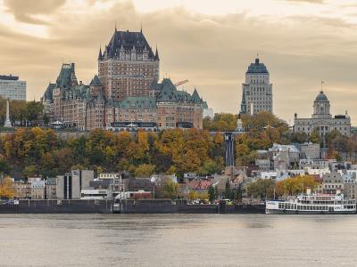 The Château Frontenac towers over Old Quebec's fortified clifftop skyline above the St. Lawrence River, with autumn foliage, the Lower Town waterfront, and a ferry at the dock as seen from Lévis.