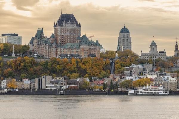 The Château Frontenac towers over Old Quebec's fortified clifftop skyline above the St. Lawrence River, with autumn foliage, the Lower Town waterfront, and a ferry at the dock as seen from Lévis.