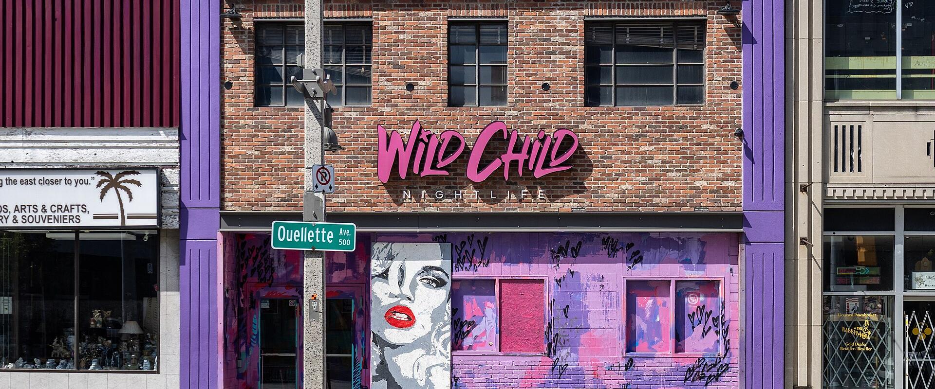 A two-story brick building with purple trim and pink signage reading "Wild Child NIGHTLIFE" on the upper facade, located at the corner of a street with a green sign for "Ouellette AVE".