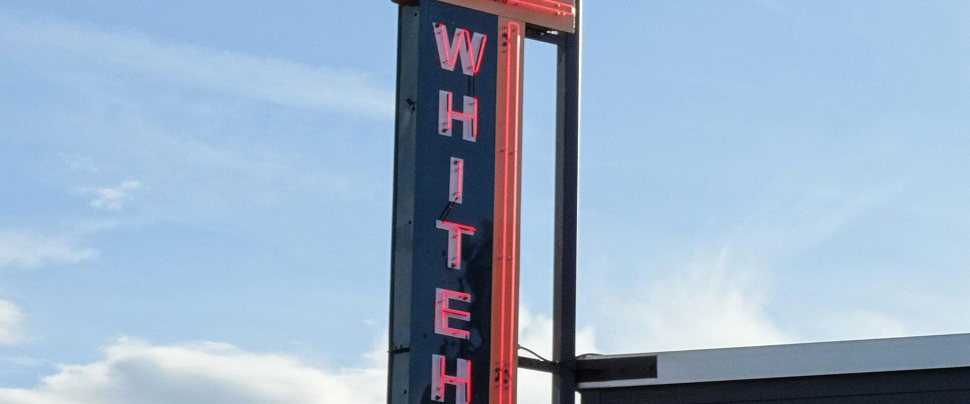 A tall vertical neon sign featuring a rearing horse silhouette at the top and the word "WHITEHORSE" written vertically in red neon letters, situated behind a wooden fence against a blue sky.