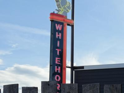 A tall vertical neon sign featuring a rearing horse silhouette at the top and the word "WHITEHORSE" written vertically in red neon letters, situated behind a wooden fence against a blue sky.