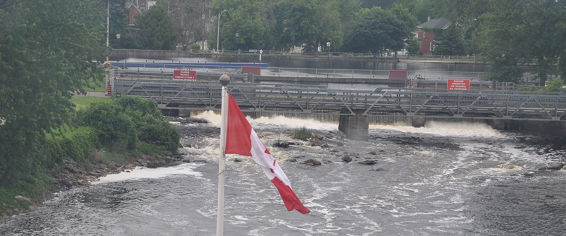 A Canadian flag flies in the foreground overlooking a river with rapids and a metal truss bridge crossing it, with red warning signs visible on the bridge structure.