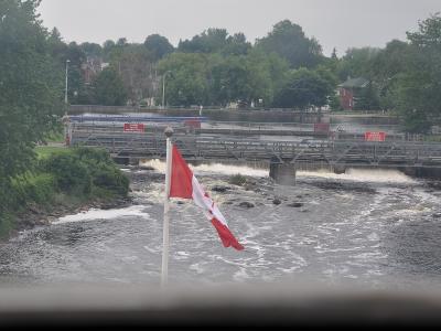 A Canadian flag flies in the foreground overlooking a river with rapids and a metal truss bridge crossing it, with red warning signs visible on the bridge structure.