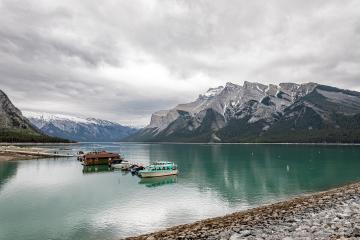 A wide shot of a turquoise alpine lake with a rocky shoreline in the foreground, featuring a floating dock with a brown building and a light blue tour boat, backed by steep, rocky mountains under a cloudy sky.