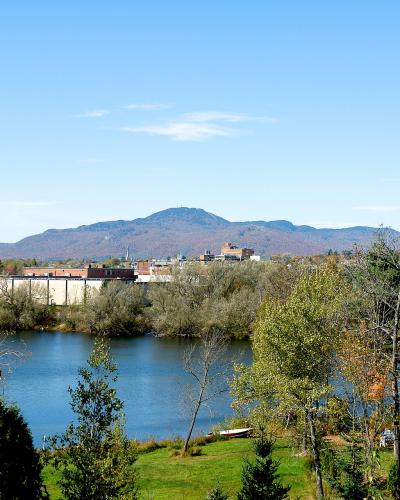 A landscape view of a river in the foreground, a town area with buildings and trees in the middle ground, and a large mountain in the background under a blue sky.