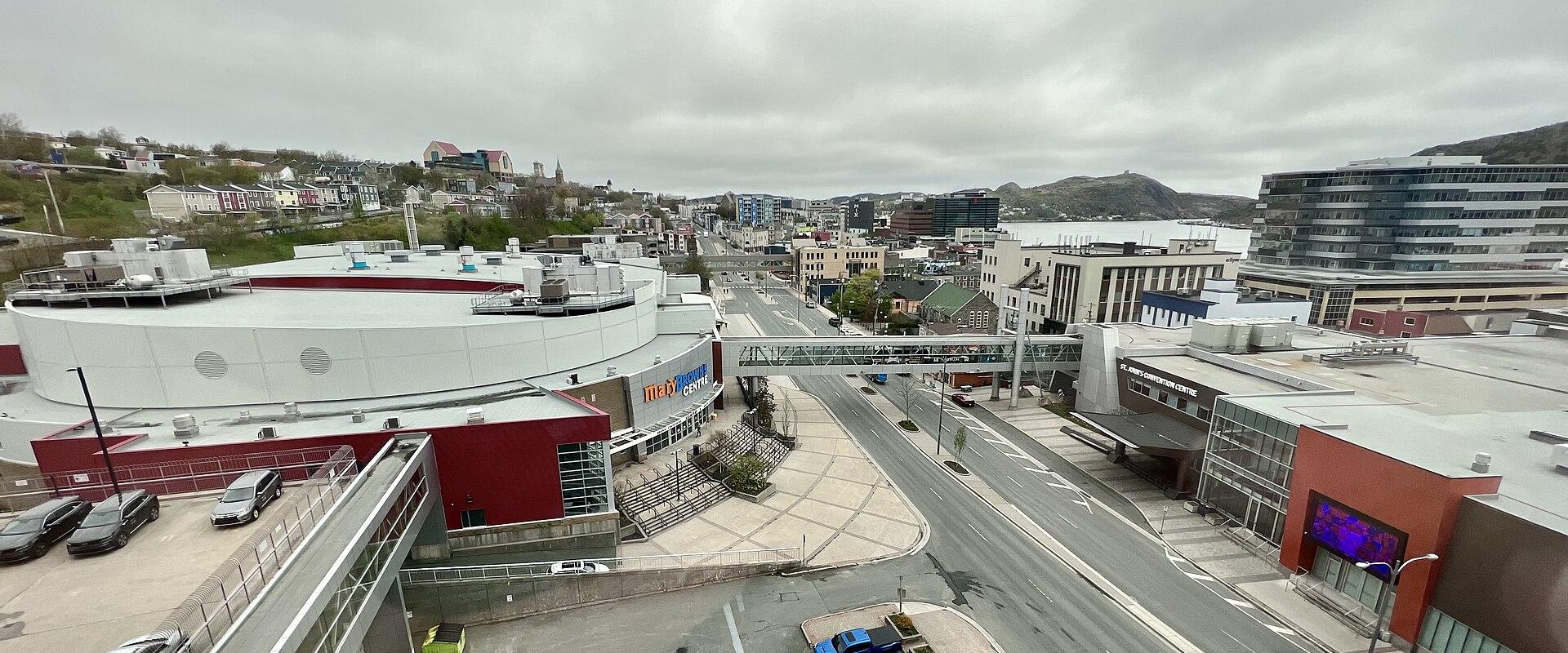 An elevated view of a large white circular arena building with red accents, situated next to a street and parking lot in an urban setting with hills in the background.
