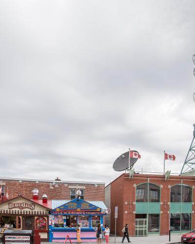 The image shows an outdoor street scene with food kiosks, including a "BeaverTails" stand and a blue "Hot N' Fries" cart, situated in front of brick buildings and a tall telecommunications tower bearing a "Bell Media" sign.