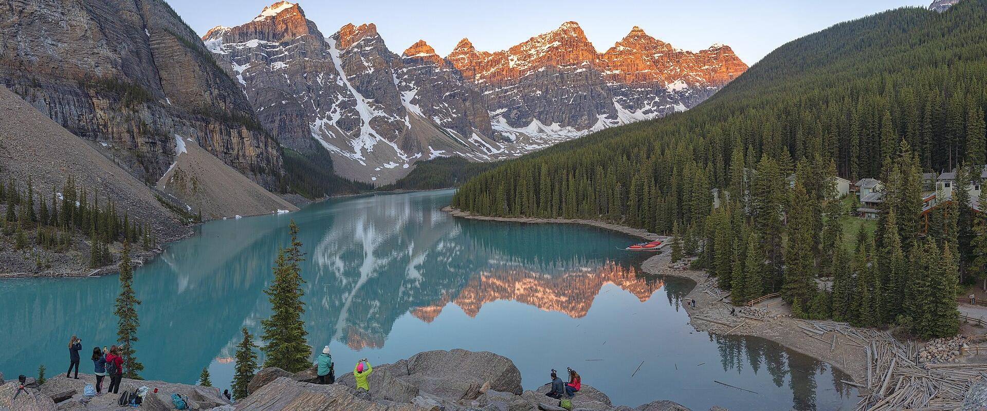 Sunrise alpenglow lights the snow-streaked Valley of the Ten Peaks above the turquoise waters of Moraine Lake in Banff National Park, Alberta, with evergreen forest and the rocky viewpoint in the foreground.