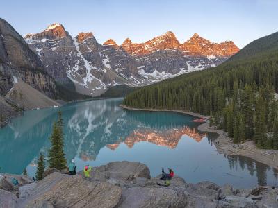 Sunrise alpenglow lights the snow-streaked Valley of the Ten Peaks above the turquoise waters of Moraine Lake in Banff National Park, Alberta, with evergreen forest and the rocky viewpoint in the foreground.