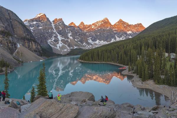 Sunrise alpenglow lights the snow-streaked Valley of the Ten Peaks above the turquoise waters of Moraine Lake in Banff National Park, Alberta, with evergreen forest and the rocky viewpoint in the foreground.