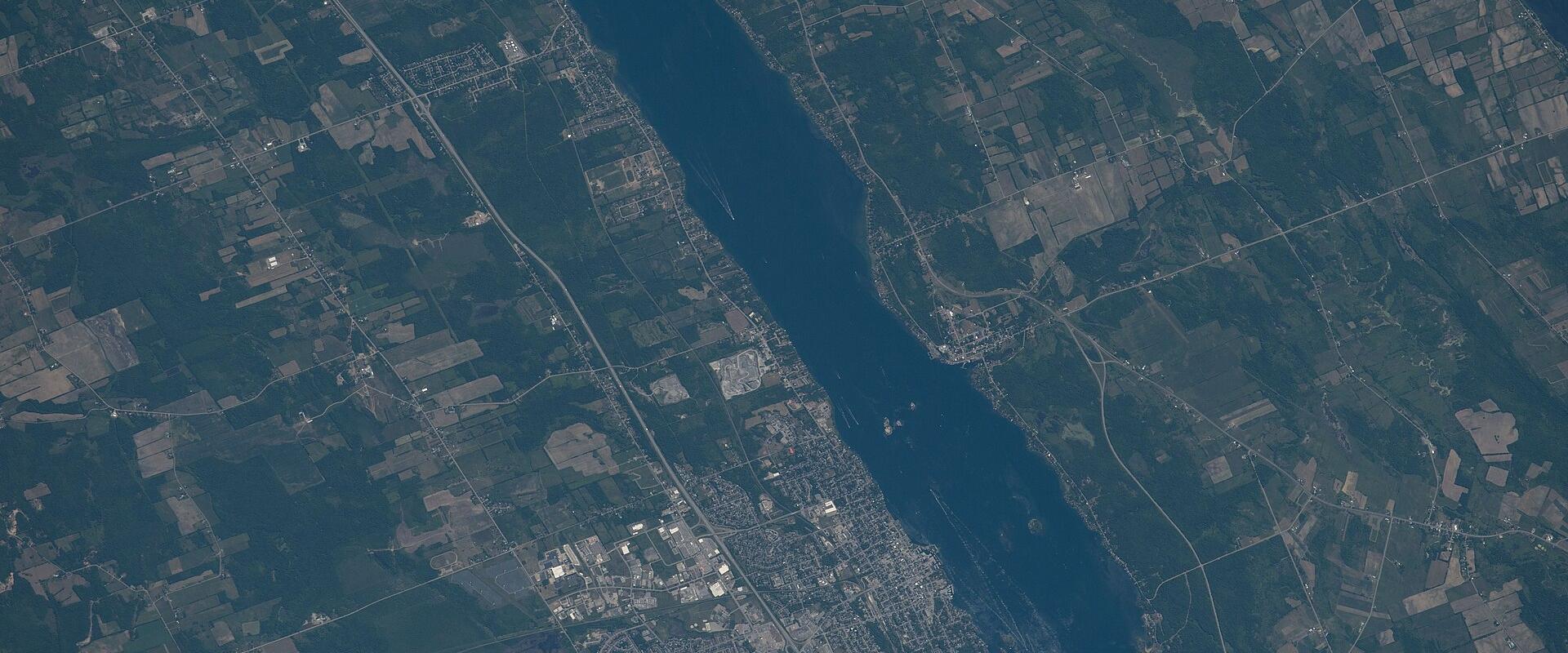 A satellite/aerial photograph showing a wide river (the St. Lawrence River) with a town situated along its banks, surrounded by agricultural fields and forested land.