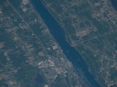 A satellite/aerial photograph showing a wide river (the St. Lawrence River) with a town situated along its banks, surrounded by agricultural fields and forested land.