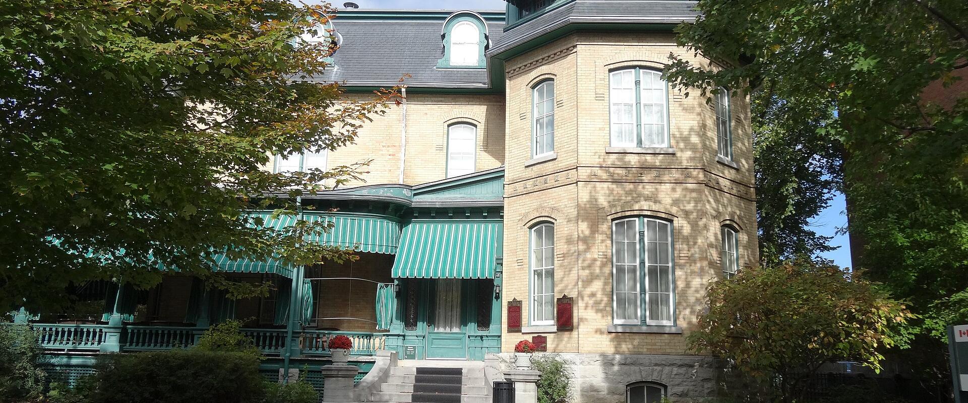 A large, light-colored brick mansion with distinctive green trim, striped awnings, and a prominent corner turret, viewed from a garden path.