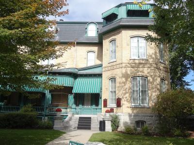 A large, light-colored brick mansion with distinctive green trim, striped awnings, and a prominent corner turret, viewed from a garden path.