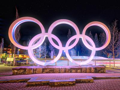 A large, illuminated sculpture of the five interlocking Olympic rings stands on a stone base in a paved public plaza at night, with buildings and trees visible in the background.