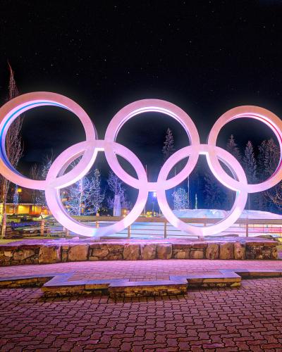 A large, illuminated sculpture of the five interlocking Olympic rings stands on a stone base in a paved public plaza at night, with buildings and trees visible in the background.