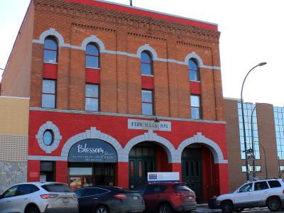 A red brick building with arched windows and garage doors, featuring a sign reading "FIRE HALL No1" and a business sign for "Blossom" below.