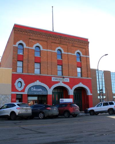 A red brick building with arched windows and garage doors, featuring a sign reading "FIRE HALL No1" and a business sign for "Blossom" below.