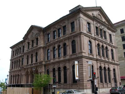 A large, ornate, multi-story sandstone building with arched windows and classical architectural details, situated on a city street corner with traffic lights and other buildings nearby.