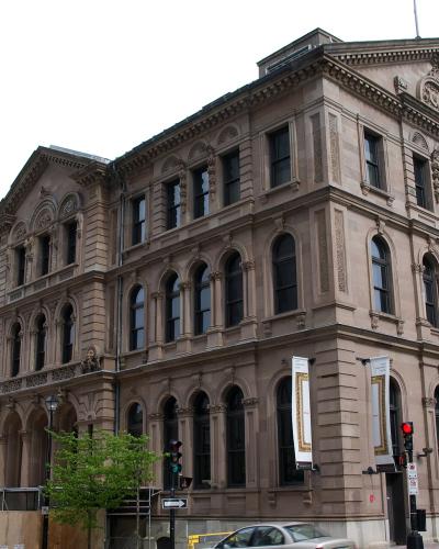 A large, ornate, multi-story sandstone building with arched windows and classical architectural details, situated on a city street corner with traffic lights and other buildings nearby.