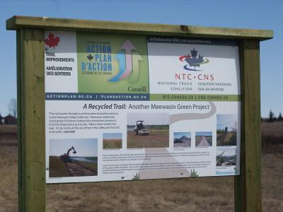 A wooden information kiosk sign displaying details about "A Recycled Trail: Another Meewasin Green Project" with logos for the National Trails Coalition and Canada's Economic Action Plan.