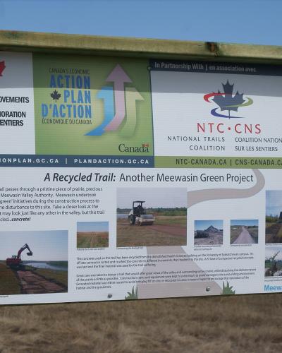 A wooden information kiosk sign displaying details about "A Recycled Trail: Another Meewasin Green Project" with logos for the National Trails Coalition and Canada's Economic Action Plan.