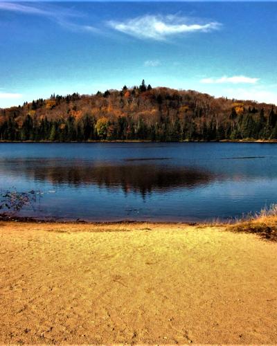 A sandy beach in the foreground leading to a calm lake, with a forested hill displaying autumn foliage in the background under a blue sky.