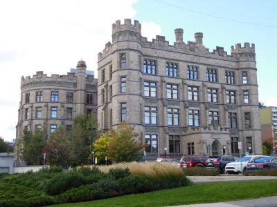 A large, grey stone building with Gothic Revival architecture featuring crenellated towers and arched windows, with a parking lot and landscaping in the foreground.