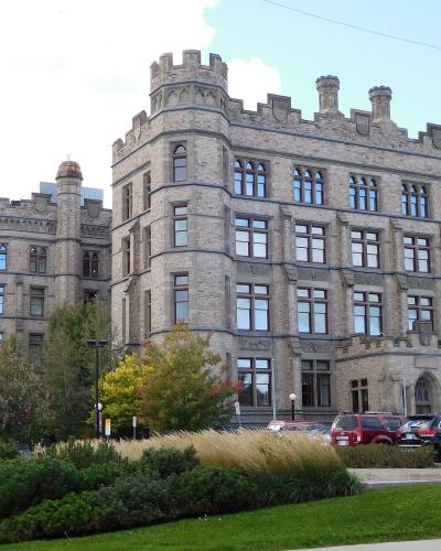 A large, grey stone building with Gothic Revival architecture featuring crenellated towers and arched windows, with a parking lot and landscaping in the foreground.