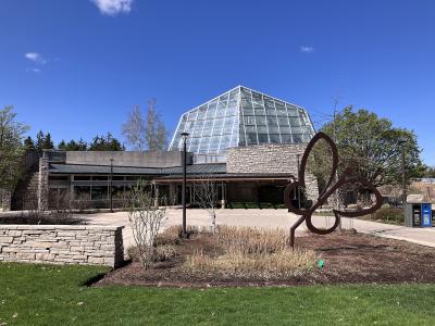 The image shows the exterior of a glass-domed conservatory building with stone walls and a large metal butterfly sculpture in the foreground.