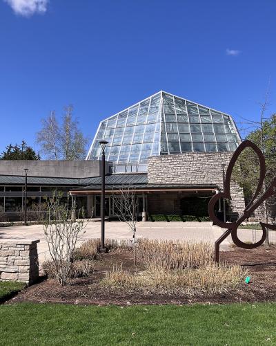 The image shows the exterior of a glass-domed conservatory building with stone walls and a large metal butterfly sculpture in the foreground.