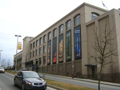 A beige, multi-story institutional building with large arched windows and vertical banners displaying natural history imagery (fossils, plants) and French text, situated next to a street with cars.