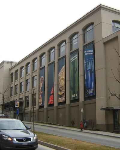 A beige, multi-story institutional building with large arched windows and vertical banners displaying natural history imagery (fossils, plants) and French text, situated next to a street with cars.